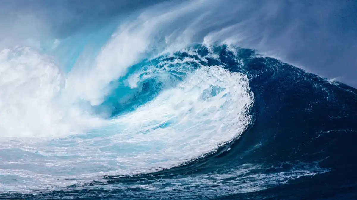A towering ocean wave curling over itself under a dark sky, with churning white foam and deep blue water.