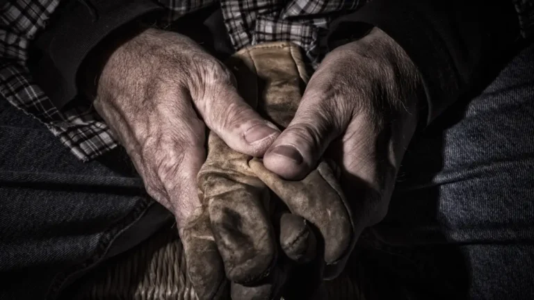 Weathered hands gripping old work gloves, resting in quiet tension.
