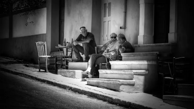 Three elderly men sit on worn steps outside a modest building, talking in the sunlit shade; a few empty chairs and a small table with bottles suggest long, slow conversation.