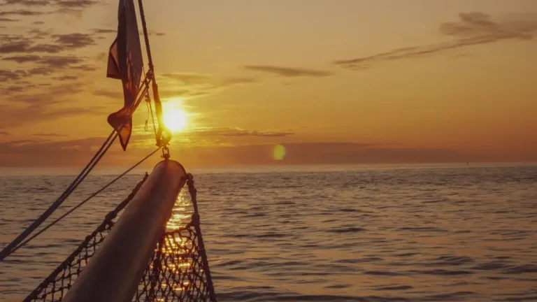 Bow of a sailing ship at sea during sunset, heading toward the horizon under an orange sky.