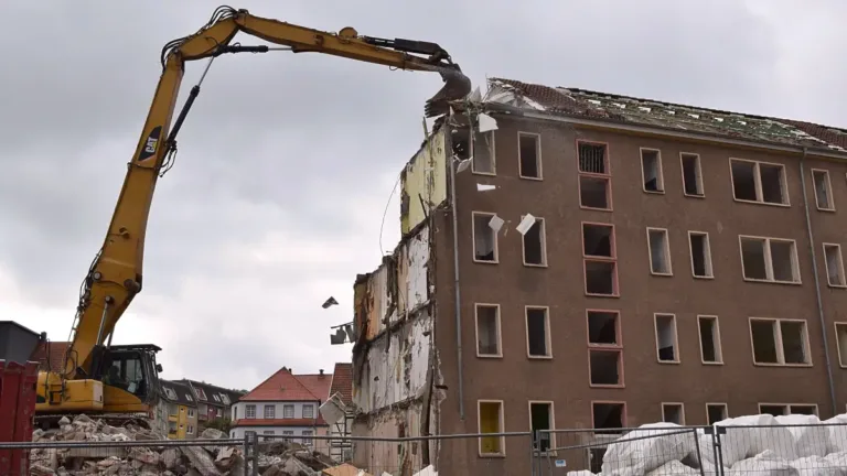 A demolition excavator tears into the top floor of a partially dismantled apartment building under grey skies.