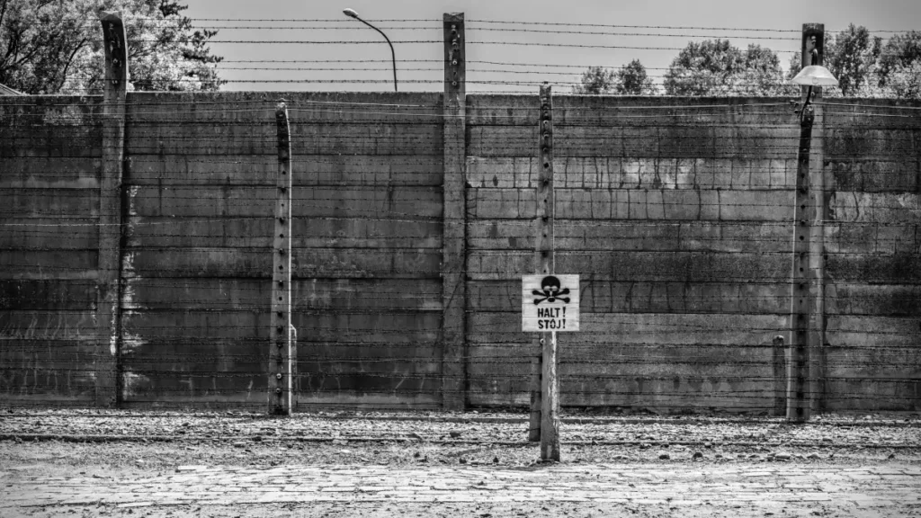 Black-and-white photograph of a barbed-wire fence and concrete wall with a warning sign reading “Halt! Stoj!” marked with a skull and crossbones.