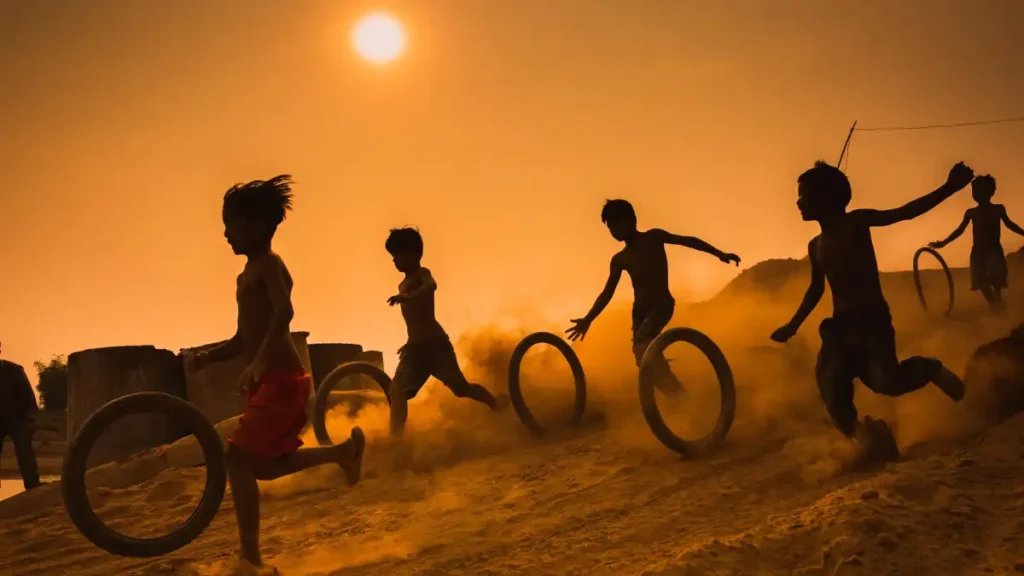 Children running barefoot at sunset, rolling tires through dust, silhouettes glowing in golden light.