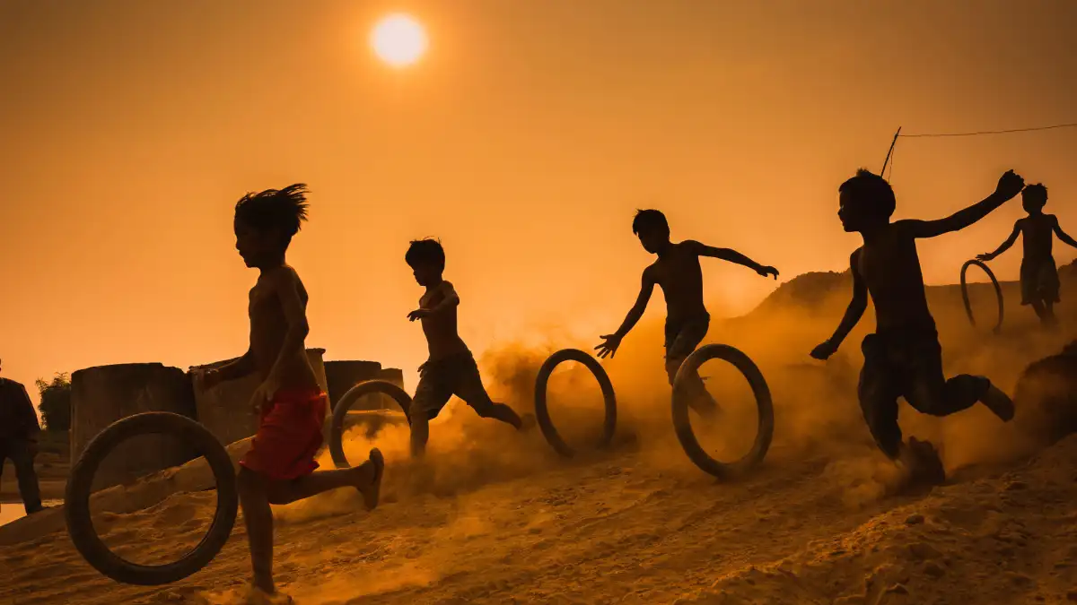 Children running barefoot at sunset, rolling tires through dust, silhouettes glowing in golden light.