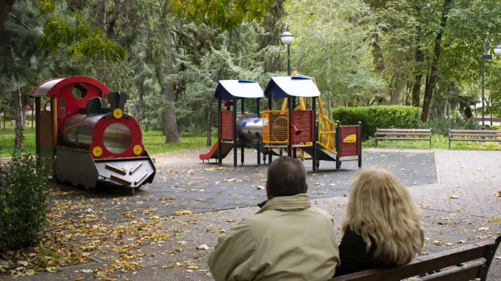 Elderly couple sitting on a park bench, watching an empty playground with a toy train and climbing structures, symbolizing declining birthrates.