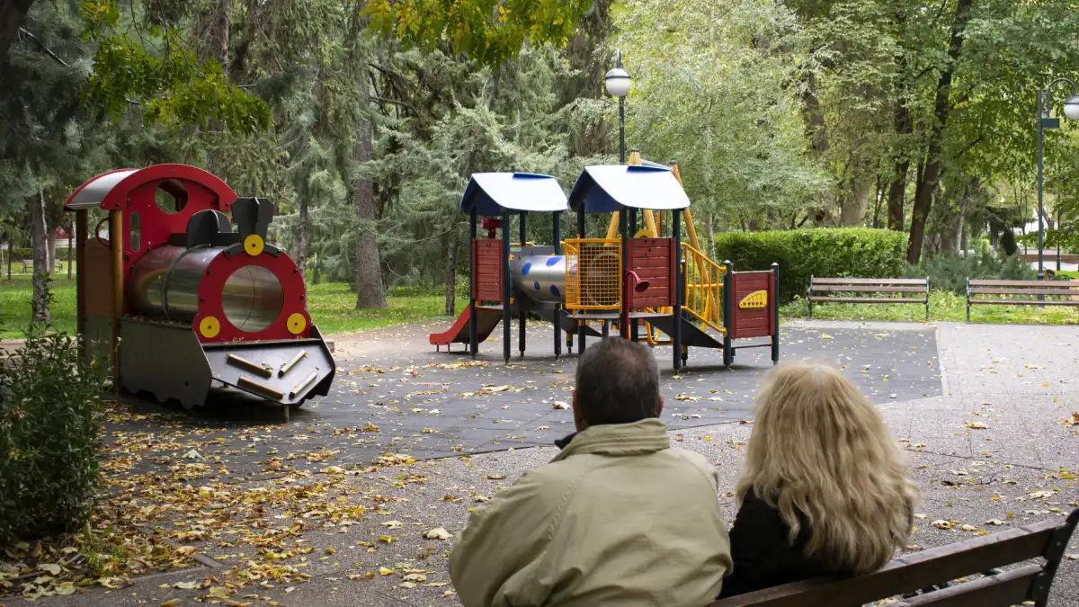 Elderly couple sitting on a park bench, watching an empty playground with a toy train and climbing structures, symbolizing declining birthrates.