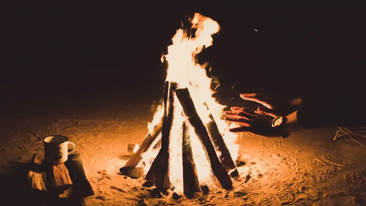 A campfire burning in the dark, with a person’s hands reaching out for warmth and a mug resting on the ground beside the fire.