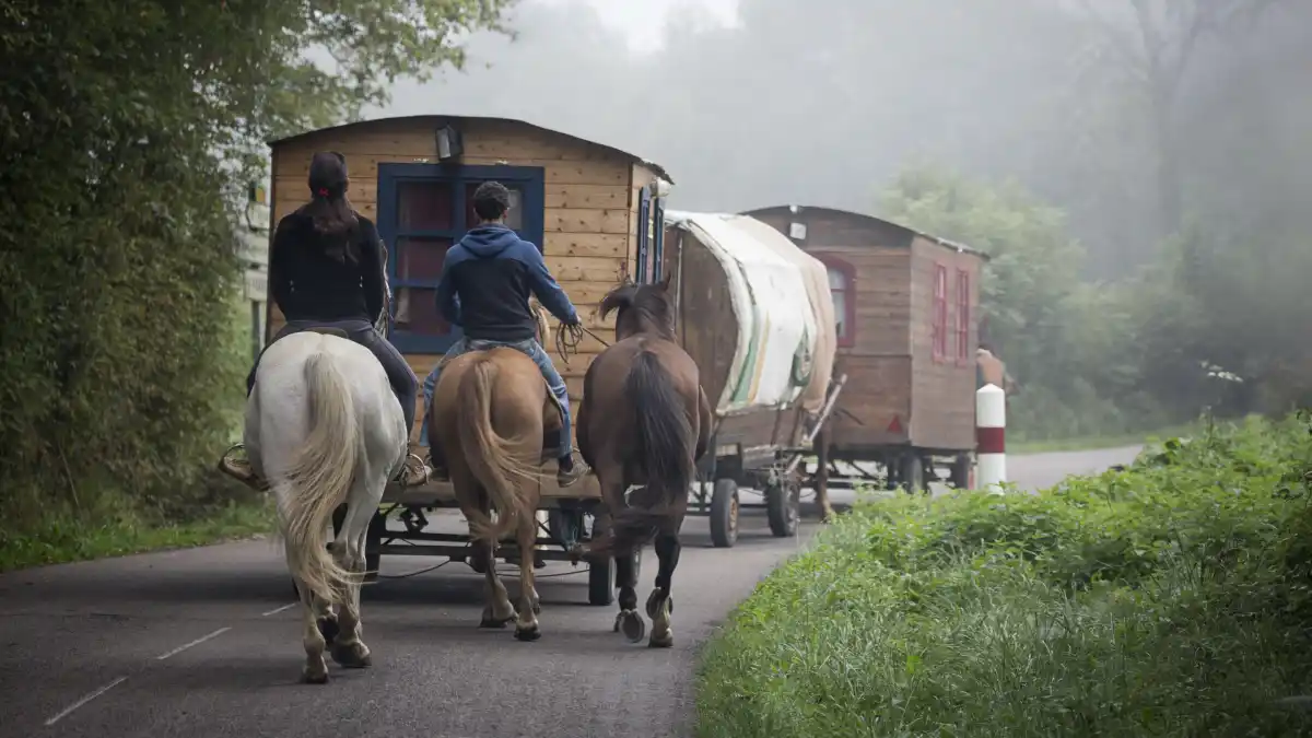 Two riders on horseback travel alongside horse-drawn wagons on a misty rural road.