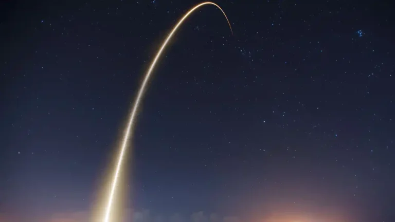 A rocket launch captured at night, leaving a bright curved trail of light across a star-filled sky.