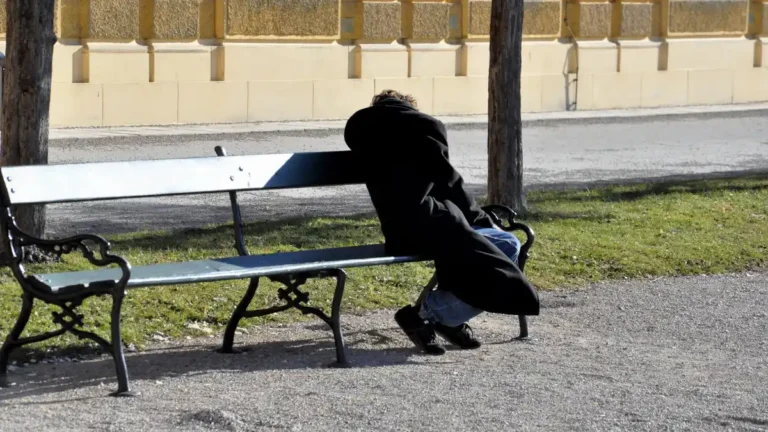 A person in a long dark coat sleeping slumped on a park bench in daylight, alone, with their face hidden, in an otherwise peaceful urban setting.