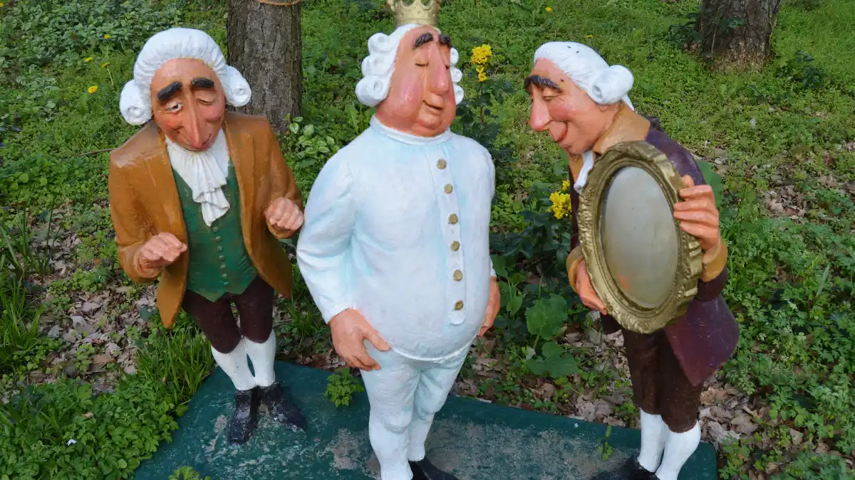 Three cartoonish statues depicting the tale of “The Emperor’s New Clothes”: a king in white undergarments and crown stands between two courtiers who admire him, one holding a mirror, set outdoors among grass and trees.