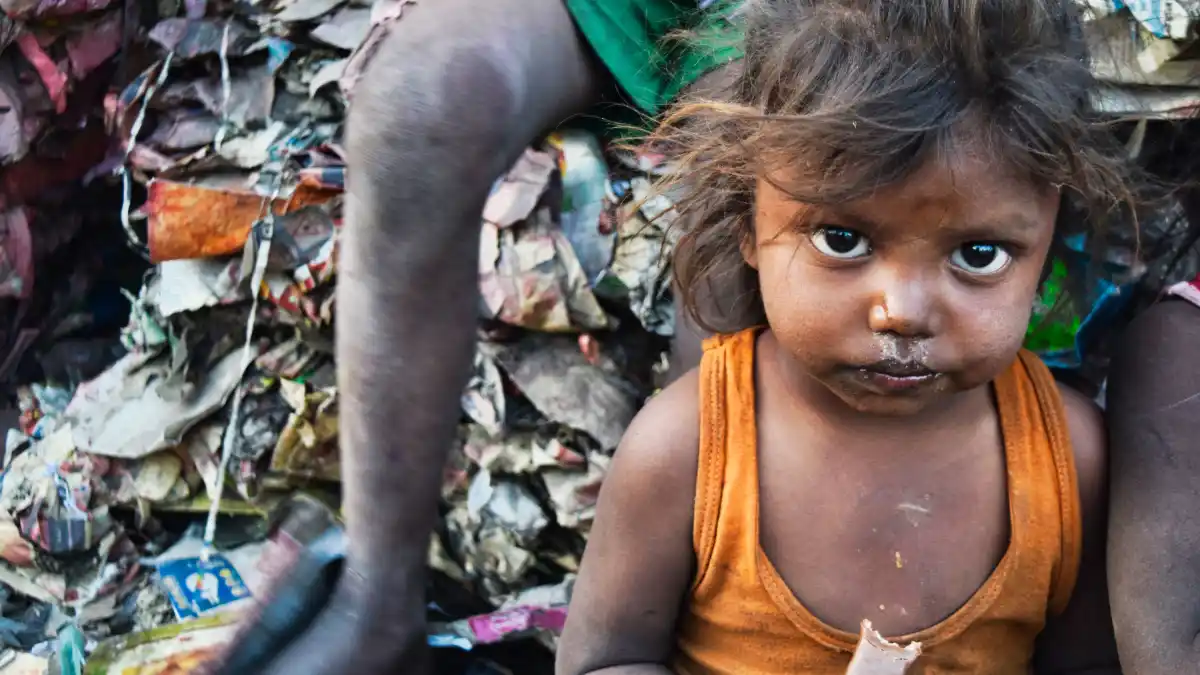 Young child sitting amid piles of waste, with dirt on face and clothing, illustrating extreme poverty and inequality.