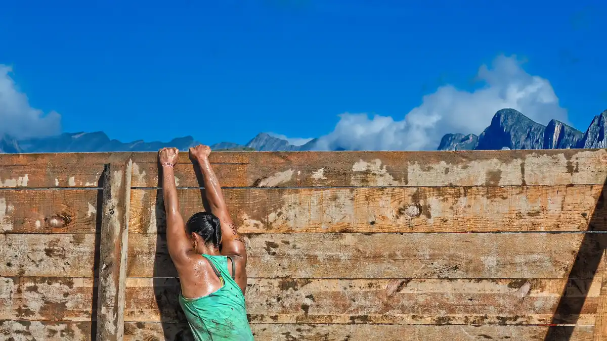 Person climbing over a weathered wooden barrier with arms raised, with rugged mountains and blue sky in the background.