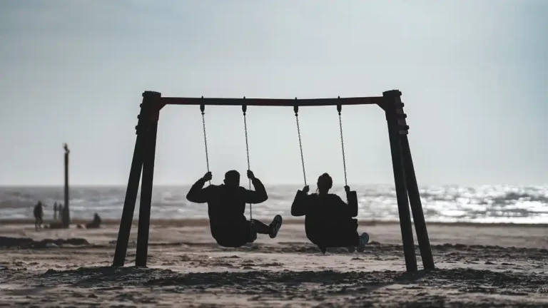 Two people on swings facing the sea on a cloudy day, silhouetted against the light reflecting off the water.