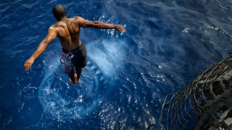 Man mid-air leaping into the deep blue ocean, captured from behind as he plunges away from a ship’s edge.