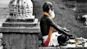 Elderly man sitting in meditation by a riverside shrine, draped in a vivid orange cloth amid a monochrome landscape — symbol of solitude and contemplation.