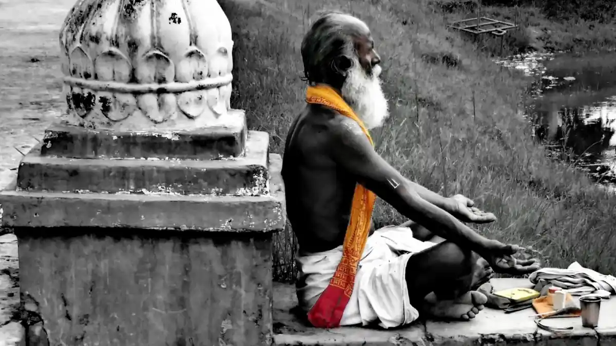 Elderly man sitting in meditation by a riverside shrine, draped in a vivid orange cloth amid a monochrome landscape — symbol of solitude and contemplation.