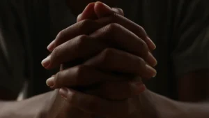 Clasped hands held together in a dark, contemplative setting, suggesting restraint, reflection, and tension rather than prayer.