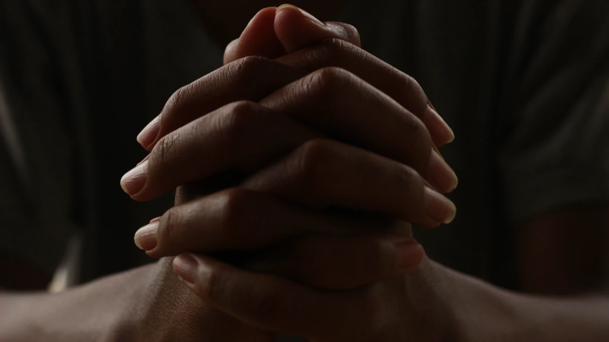 Clasped hands held together in a dark, contemplative setting, suggesting restraint, reflection, and tension rather than prayer.