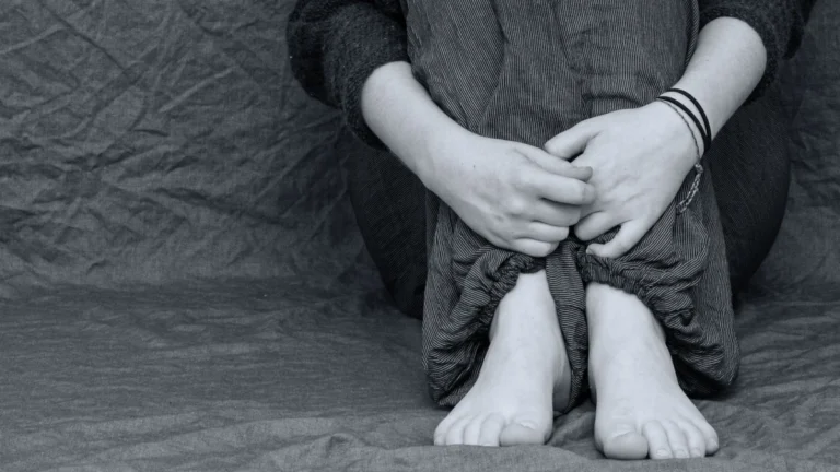 A person sitting on the floor with knees drawn to the chest, hands clasped around their legs, barefoot, conveying quiet confinement and withdrawal.