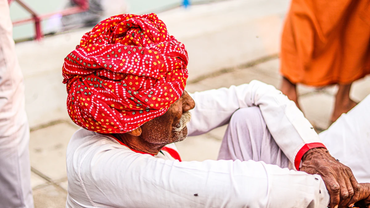 Elderly Indian man wearing a red turban and white clothing, seated outdoors with arms resting on his knees