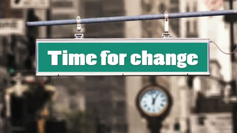 Green street sign reading “Time for change” suspended above a blurred urban street with a clock visible in the background.