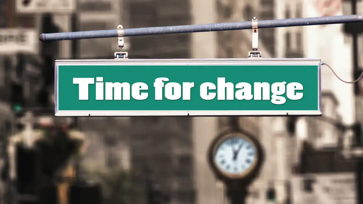 Green street sign reading “Time for change” suspended above a blurred urban street with a clock visible in the background.