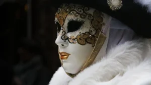 Ornate white Venetian carnival mask with gold detailing and feathers, symbolizing the hidden identities and polite illusions behind social and institutional roles.
