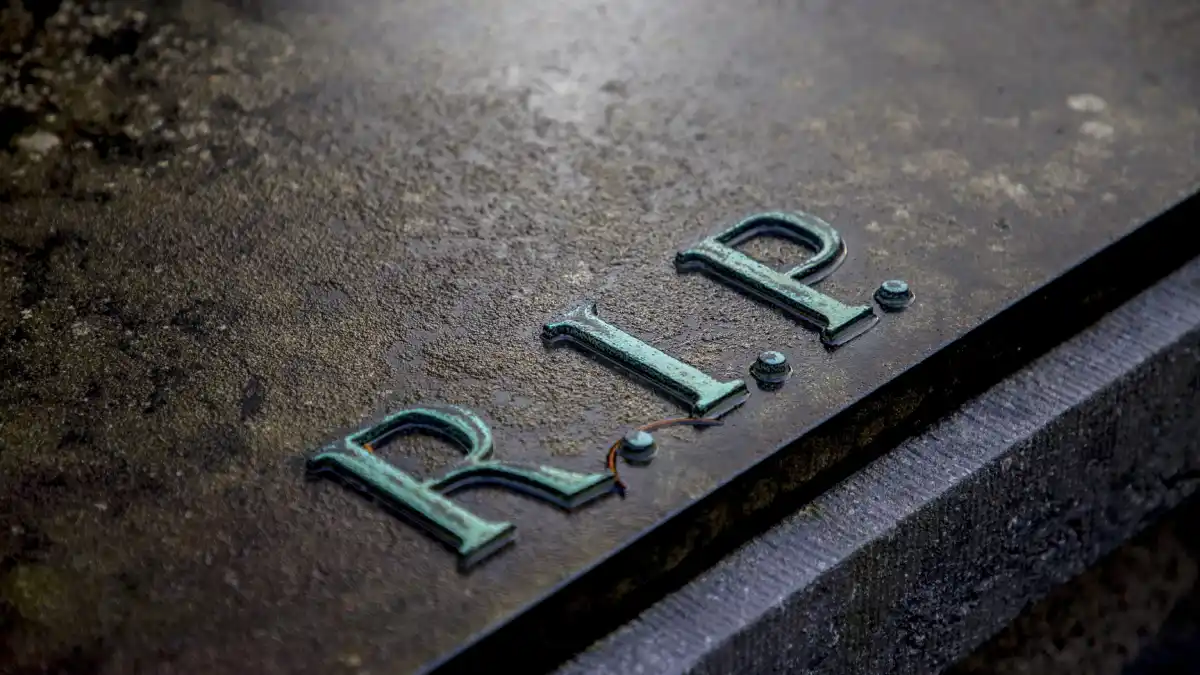 Close-up photograph of a weathered gravestone with the letters "R.I.P." engraved on its surface, viewed at an angle in low natural light.