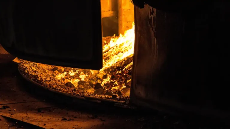 A dark industrial furnace with an open firebox glowing intensely with flames and burning embers, surrounded by heavy black metal and deep shadows.