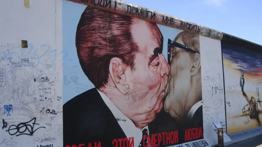 A large political mural depicts two suited male figures kissing against a graffiti-covered wall, with additional painted imagery extending along the wall beneath a clear blue sky.