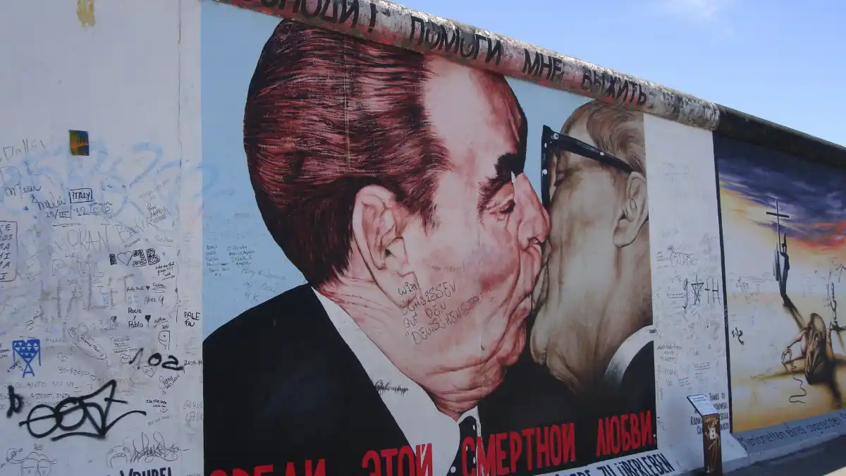 A large political mural depicts two suited male figures kissing against a graffiti-covered wall, with additional painted imagery extending along the wall beneath a clear blue sky.