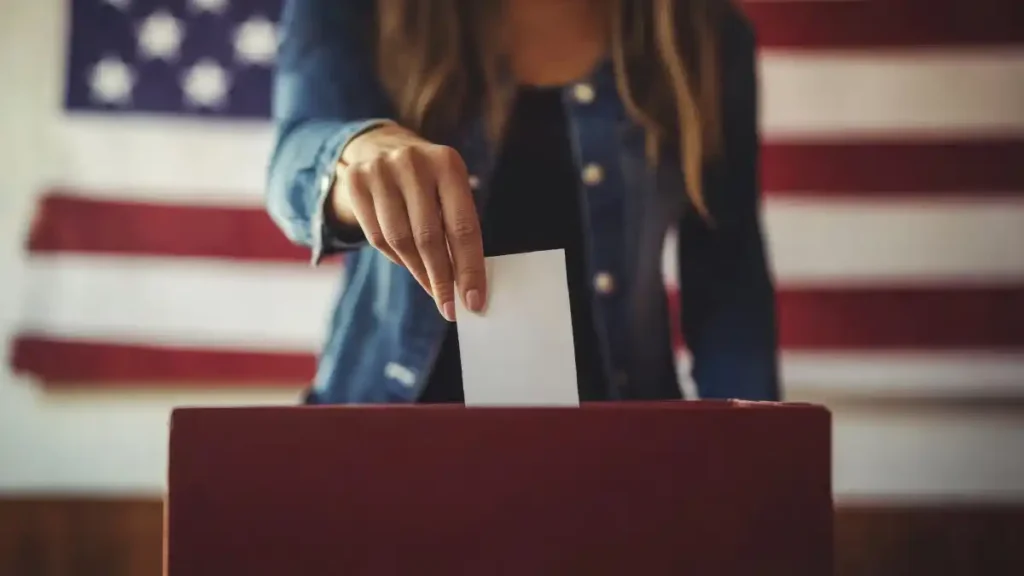 A person placing a ballot into a ballot box in front of an American flag.