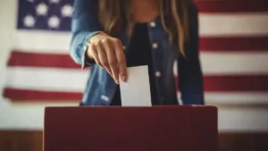 A person placing a ballot into a ballot box in front of an American flag.