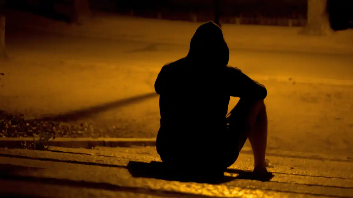 A hooded figure sits alone on a pavement at night, silhouetted against the amber glow of a streetlight.